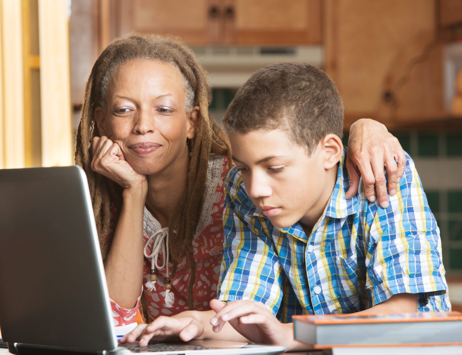 Mother overlooks as her teenaged son uses his laptop to study in their kitchen.