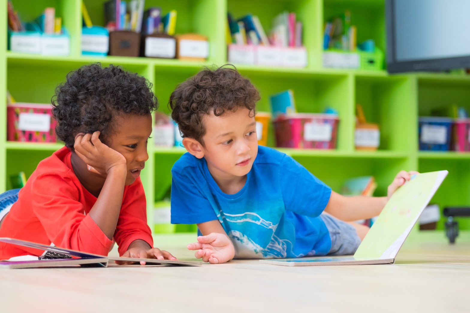 Two boys laying down on floor and reading a book in the preschool library.